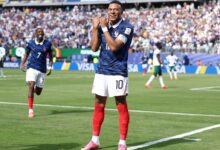 Kylian Mbappé celebra un gol con los brazos cruzados sobre el pecho durante un partido del Mundial 2026. Viste el uniforme de Francia con el número 10. Al fondo, Ousmane Dembélé corre a festejar con él en un estadio lleno.