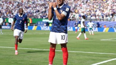 Kylian Mbappé celebra un gol con los brazos cruzados sobre el pecho durante un partido del Mundial 2026. Viste el uniforme de Francia con el número 10. Al fondo, Ousmane Dembélé corre a festejar con él en un estadio lleno.