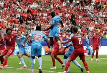 El delantero congoleño Yoane Wissa salta imponente en el área para conectar un cabezazo ante la mirada de defensores portugueses como Renato Veiga (13) y Bruno Fernandes (8). Al fondo, Cristiano Ronaldo observa la jugada desde la banda ante un estadio repleto.