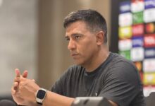 Un plano medio lateral muestra al director técnico de fútbol venezolano César Farías sentado frente a una mesa en una sala de prensa, mirando fijamente hacia la derecha con expresión seria y concentrada.