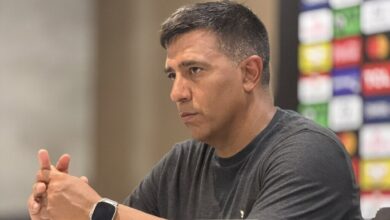 Un plano medio lateral muestra al director técnico de fútbol venezolano César Farías sentado frente a una mesa en una sala de prensa, mirando fijamente hacia la derecha con expresión seria y concentrada.