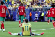 El futbolista brasileño Raphinha, de espaldas y sentado sobre el césped con las piernas completamente estiradas, refleja frustración tras el partido contra Marruecos. Al fondo se observa a los jugadores marroquíes Chadi Riad (18) e Issa Diop (14) celebrando o caminando por el campo de juego.