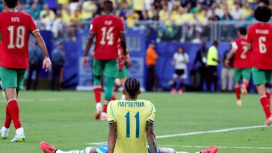 El futbolista brasileño Raphinha, de espaldas y sentado sobre el césped con las piernas completamente estiradas, refleja frustración tras el partido contra Marruecos. Al fondo se observa a los jugadores marroquíes Chadi Riad (18) e Issa Diop (14) celebrando o caminando por el campo de juego.