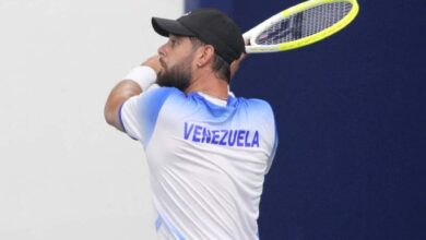 Tenista del equipo de Venezuela ejecutando un golpe de revés a dos manos durante un partido de la Copa Davis. Viste una camiseta blanca con degradado azul en los hombros, gorra negra y sostiene una raqueta con marco de color amarillo fluorescente.