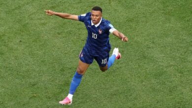 El futbolista francés Kylian Mbappé, vistiendo el uniforme azul de su selección con el número 10, corre sonriente sobre el césped verde con los brazos extendidos para celebrar un gol.