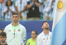 El portero Emiliano "Dibu" Martínez mira al frente con seriedad mientras Lionel Messi levanta la cabeza con los ojos cerrados durante un partido de la selección argentina.