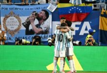 El futbolista Lionel Messi con el uniforme número 10 de la Selección de Argentina abraza a su compañero Cristian "Cuti" Romero en la cancha durante un partido del Mundial 2026. Al fondo, la tribuna muestra banderas argentinas, una bandera de Venezuela y una pancarta de homenaje a Messi.