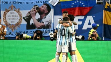 El futbolista Lionel Messi con el uniforme número 10 de la Selección de Argentina abraza a su compañero Cristian "Cuti" Romero en la cancha durante un partido del Mundial 2026. Al fondo, la tribuna muestra banderas argentinas, una bandera de Venezuela y una pancarta de homenaje a Messi.
