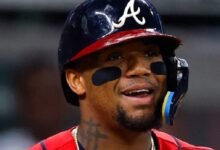 Primer plano del pelotero venezolano Ronald Acuña Jr. sonriendo con el uniforme de los Bravos de Atlanta, usando su casco de bateo con la letra 'A' y parches negros bajo los ojos.