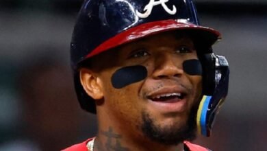 Primer plano del pelotero venezolano Ronald Acuña Jr. sonriendo con el uniforme de los Bravos de Atlanta, usando su casco de bateo con la letra 'A' y parches negros bajo los ojos.