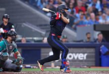 Andrés Giménez completa un swing con el bate en el home plate. El pelotero lleva el uniforme alternativo oscuro de los Toronto Blue Jays, con detalles en azul brillante y rojo, mientras el catcher de los Seattle Mariners sigue la jugada.