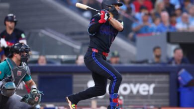 Andrés Giménez completa un swing con el bate en el home plate. El pelotero lleva el uniforme alternativo oscuro de los Toronto Blue Jays, con detalles en azul brillante y rojo, mientras el catcher de los Seattle Mariners sigue la jugada.