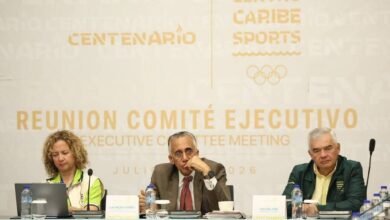 Reunión del Comité Ejecutivo de Centro Caribe Sports. En el centro, el presidente de la organización, Luis Mejía Oviedo, habla frente a un micrófono flanqueado por dos directivos en una mesa de conferencia con identificadores y botellas de agua.