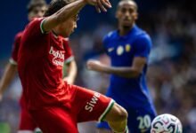 El futbolista venezolano Jon Aramburu, vistiendo el uniforme rojo de la Real Sociedad, remata de volea con la pierna izquierda un balón en el aire durante el partido de pretemporada contra el Chelsea en Stamford Bridge. Al fondo, se observa al jugador del Chelsea Axel Disasi con la camiseta azul número 20 siguiendo la jugada.