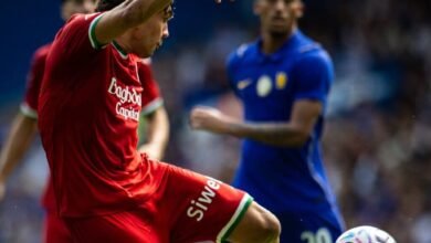El futbolista venezolano Jon Aramburu, vistiendo el uniforme rojo de la Real Sociedad, remata de volea con la pierna izquierda un balón en el aire durante el partido de pretemporada contra el Chelsea en Stamford Bridge. Al fondo, se observa al jugador del Chelsea Axel Disasi con la camiseta azul número 20 siguiendo la jugada.