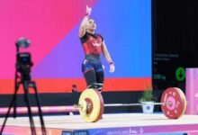 Fotografía de la pesista venezolana Katherin Echandía celebrando sobre la tarima de levantamiento de pesas. Viste un traje de halterofilia en colores rojo, azul y negro con la palabra "Ven" en el pecho. La atleta levanta el brazo derecho apuntando hacia el cielo en señal de victoria frente a una barra cargada con discos Eleiko de color amarillo y rojo.