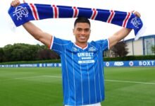 Kevin Kelsy sonríe mientras sostiene una bufanda azul y blanca sobre su cabeza con ambas manos. Viste una camiseta de fútbol azul de rayas diplomáticas blancas finas con el logotipo de Unibet en el centro. El fondo muestra un campo de fútbol verde bajo un cielo ligeramente nublado.