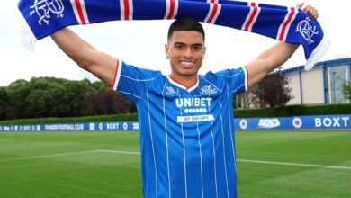 Kevin Kelsy sonríe mientras sostiene una bufanda azul y blanca sobre su cabeza con ambas manos. Viste una camiseta de fútbol azul de rayas diplomáticas blancas finas con el logotipo de Unibet en el centro. El fondo muestra un campo de fútbol verde bajo un cielo ligeramente nublado.