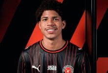 Un primer plano del futbolista venezolano David Martínez sonriendo con una camiseta de fútbol de color negro con finas líneas rojas verticales, que lleva los logotipos de Puma, Jack & Jones y el escudo del FC Midtjylland, con un fondo de franjas diagonales negras y rojas