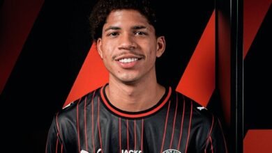 Un primer plano del futbolista venezolano David Martínez sonriendo con una camiseta de fútbol de color negro con finas líneas rojas verticales, que lleva los logotipos de Puma, Jack & Jones y el escudo del FC Midtjylland, con un fondo de franjas diagonales negras y rojas