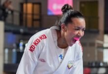 Un plano medio de la karateca venezolana Yorgelis Salazar en pleno tatami, capturada de perfil con una expresión de máxima euforia y garra tras asegurar una victoria. La atleta, que lleva el cabello recogido en un moño alto y un protector bucal rosado, grita efusivamente con la boca abierta. Viste su karategui blanco oficial, donde destacan las letras rojas de la marca patrocinadora en el cuello y el escudo del Comité Olímpico Venezolano bordado en el pecho.