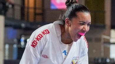 Un plano medio de la karateca venezolana Yorgelis Salazar en pleno tatami, capturada de perfil con una expresión de máxima euforia y garra tras asegurar una victoria. La atleta, que lleva el cabello recogido en un moño alto y un protector bucal rosado, grita efusivamente con la boca abierta. Viste su karategui blanco oficial, donde destacan las letras rojas de la marca patrocinadora en el cuello y el escudo del Comité Olímpico Venezolano bordado en el pecho.