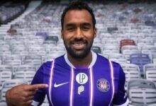 El mediocampista venezolano Cristian Cásseres Jr. sonríe vistiendo la camiseta morada y blanca con rayas verticales del Toulouse FC, mientras señala con su dedo el escudo del club francés en un estadio vacío.