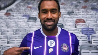 El mediocampista venezolano Cristian Cásseres Jr. sonríe vistiendo la camiseta morada y blanca con rayas verticales del Toulouse FC, mientras señala con su dedo el escudo del club francés en un estadio vacío.