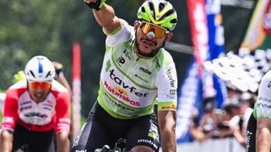 El ciclista venezolano Leangel Linarez celebra con el puño derecho en alto al ganar una etapa de la Vuelta a Venezuela, vistiendo el uniforme verde y blanco del equipo Tavfer-Ovos Matinados-Mortágua, con gafas de sol doradas y casco verde fosforescente, mientras otros competidores aparecen detrás